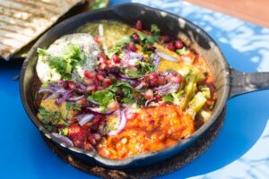 Shaksouka Hola Mexico. Eggs baked in aromatic tomato and peppers paste, with beans, served with marinated cactus, guacamole, salsa verde, salsa chipotle, cheddar and tortillas