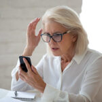 Confused,Senior,Businesswoman,Sit,At,Office,Desk,Hold,Cellphone,Experience